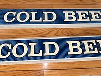 Close-up photo of two plastic Budweiser beer signs side by side on a wooden floor, showing 'COLD BEER' text