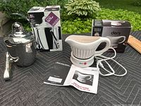 Oneida stainless steel ice bucket with lid and matching tongs next to its original box, alongside a white ceramic Deni electric gravy warmer, instruction manuals, and its box all displayed on black quilted surface outdoors with green foliage background.