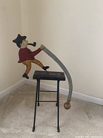 Full view of the metal folk art sawman figurine standing on a black stool with a curved saw blade and weighted pendulum.