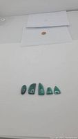 Five green malachite cabochon gemstones displayed horizontally on white surface with two envelopes and a coin in background for scale.