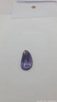 Polished natural charoite gemstone teardrop shape, showing purple coloration with dark and light patterns.