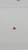 Close-up photo of a small oval-shaped natural ruby with a pinkish-red hue placed on a white background.