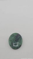 Oval cabochon ruby zoisite gemstone on flat surface showing natural green and black colors with subtle ruby inclusions.