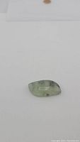 Photo of single cabochon-cut rutilated prehnite gemstone against white background, highlighting semi-translucent light green color and inclusion structure.