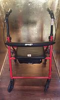 Front view of red Drive foldable walker showing padded black seat, small black storage bag under seat, and front wheels.