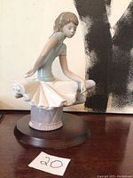 Side view showing ballerina seated with arms behind, wearing light blue top and white skirt on dark wooden base