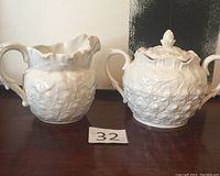 Milk dispenser and sugar bowl with lid placed side by side on a wooden surface, porcelain with textured floral pattern and handles visible, milk dispenser's repaired spout visible