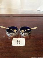 Two sterling silver salt cellars with blue glass liners and two small sterling silver spoons on a wooden surface.