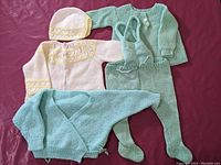 Three hand-knit infant outfits displayed on a pink background: pale teal wrap-over sweater, white and yellow button-up sweater with matching bonnet, and light green footed overalls with pullover sweater.
