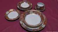 Four hand-painted Satsuma porcelain pieces including dinner plate, smaller plate, bread plate, teacup with lithophane, and saucer, showing the set's detailed color and gold trim.