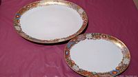 Photo showing the two oval Satsuma porcelain serving platters placed side by side on a pink surface, capturing the size difference and hand-painted floral and gold border details.