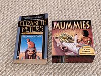 Paperback mystery novel 'The Mummy Case' and closed educational 'Mummies' craft kit box placed side by side on carpeted floor.