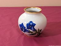Front angle of small rounded white vase with blue flowers and gold rim on top and base.