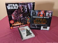 Three items shown: Star Wars Trivia Game box featuring Darth Vader, Obsessed with Star Wars electronic trivia game booklet and device, and an unopened pack of Darth Vader and Son postcards.