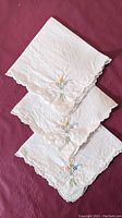 Three folded vintage white linen tea napkins with hand embroidery and scalloped edges stacked on maroon fabric background