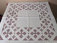 Full view of the vintage white square table linen with geometric Hardanger embroidery patterns arranged around a central plain linen square, laid flat on red surface.