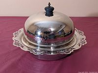 Closed view showing high-domed shiny silver lid with finial on top covering deep dish with cutout rim.