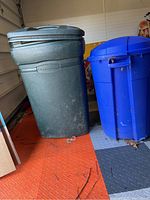 Photo showing a large green Rubbermaid garbage can and a large blue garbage can, both with lids, standing on a textured floor inside a garage. The green can shows scuffs and dirt.