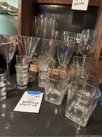Group of assorted clear bar glassware including wine glasses, shot glasses, pampas style short glasses, and tall beer pilsner glasses arranged on and near wooden shelf