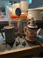 Five assorted beer steins including two with lids, arranged on a table with background items