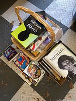 Overview of basket filled with assorted books, games, DVDs, and CDs on floor with checkered mats beneath. Wicker basket tightly packed with media items and games.