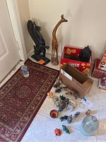 Overview of decorative animal figurines on floor, tall wooden giraffe, black cast-iron griffin statue near wall, and large clear glass bottle on floor