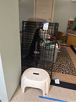 Photo showing black metal wire dog crate and white plastic stepping stool placed on carpeted floor in room corner.