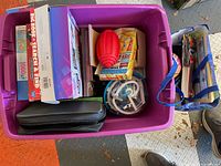 Top-down view of purple plastic tub containing boxed games, toys, a CD case, and some books.