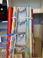 Four large rolls of wrapping paper bundled upright with straps, standing on carpet in front of white shelving