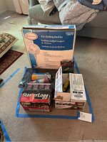 FeatureComforts brand 16 inch oscillating stand fan in original box with plastic wrapping, assorted boxes containing fire starter logs, a propane hose, paint tray, and aerosol paints arranged together on floor.