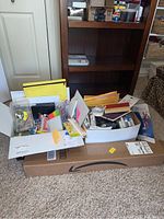 Photo showing multiple office and craft items including writing instruments, paper, and organizers placed in two white boxes and a large Amazon shipping box beneath them.