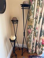 Three black metal floor candleholders of varying heights, each holding a round candle. Shown against wall and floral curtains.