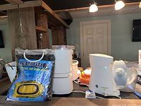 Photo showing SurfGear inflatable boat in original packaging, white Oster ice cream maker, Rival crock pot, and other kitchen and outdoor items on a table in basement room