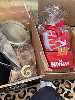 Photo showing cardboard boxes containing Wawa branded insulated cups in red carrying pouch, Wawa t-shirt, blanket, unknown coupons, and nearby smaller metal tray, decorative glass sphere, and other small items.