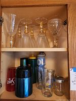 Top and middle shelves of cabinet showing 8 bubbled water glasses and 2 wine glasses with gold accents, plus a variety of other drink containers and glasses on the shelf below.