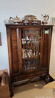 Front view of antique wooden china cupboard with glass door, showing dishes inside and silver tea sets on top