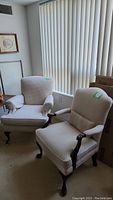 Pair of cream-colored upholstered arm chairs with small dark pattern, wooden legs and arms, sitting in front of window blinds.