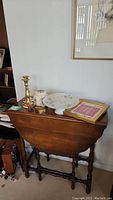 Wooden collapsible drop-leaf table with decorative items on top including brass candlesticks, ceramic jar, pedestal plate, vintage brass box, and a framed item.
