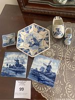 Set of six Delft blue and white ceramic items placed on a table with lace tablecloth, two square windmill tiles, a hexagonal dish with windmill design, two ceramic Dutch shoe figurines painted in Delft style, and a small square dish, all featuring traditional Dutch blue and white patterns.