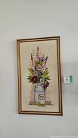 Framed embroidered artwork showing floral bouquet in a milk jug, hung on white wall.