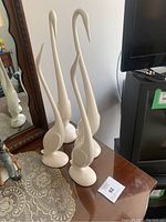 Four white mid-century ceramic crane bird sculptures displayed on a wooden side table next to a mirror and television.