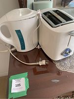 Photo showing a white electric kettle and a white 2-slot toaster placed side by side on a wooden surface, both with power cords visible.