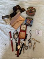 Shows full collection of various wristwatches, clocks, and a wristwatch bracelet in box laid out on white textured cloth.