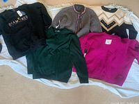 Lot of 5 women's medium sweaters laid out on a beige carpet with a white fringed throw underneath.