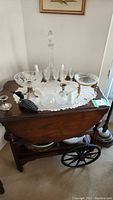 Wood bar cart topped with a white doily holding a glass decanter, matching glass bowls, salt and pepper shakers, a ceramic bird figurine, and a glass bowl with brass legs. Lower shelf holds plates and a bowl.