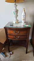Single wood bedside table with two drawers, glass top, and brass handles placed on carpeted floor with a lamp and lotion bottle on top.