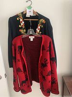 Two wool sweaters hanging on hangers shown from the front. One black with embroidered floral design and one red with black animal print.