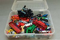 Top view of transparent plastic box filled with assorted vintage die-cast toy vehicles including Batman motorcycle figure, cars, buses, and trucks.