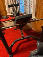 Side view of MWB 765 exercise bench showing padded backrest, metal frame, and leg support rollers with added weight plates on adjacent bench