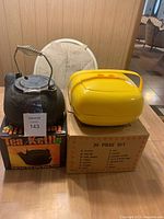 Two cast iron tea kettles and a yellow plastic container with dish set on top of its cardboard box on wooden floor.